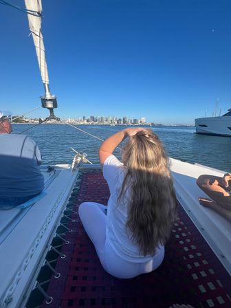 Relaxed passenger with long blonde hair sitting on a catamaran trampoline, gazing at the sunny San Diego skyline, blue harbor waters, and nearby yachts.