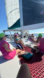 Sunlit group lounging on a white catamaran's netted deck under full sails, bright swimwear and neon caps, coastal waters and distant shoreline in the background.