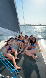 Six women in colorful bikinis and sunglasses lounging on a catamaran trampoline, holding drinks and smiling while sailing on a sunny coastal bay with sparkling water and a palm-lined shoreline.