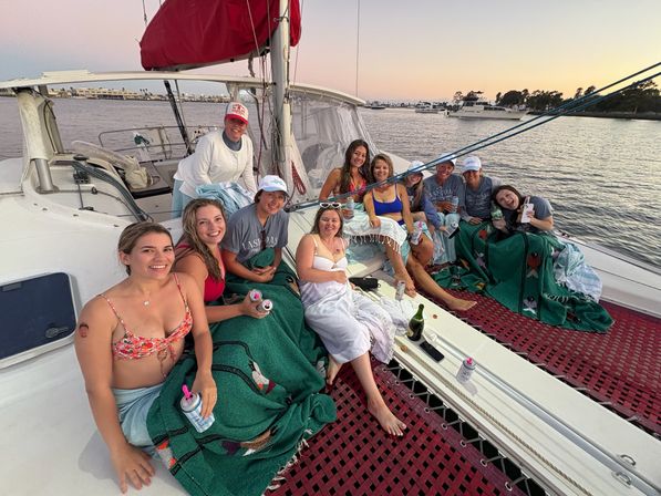 Smiling group of friends relaxing on a sailboat at sunset in a marina, wrapped in blankets on the deck netting with drinks.