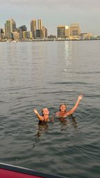 Two swimmers waving in a calm harbor at sunset, San Diego skyline and waterfront lights reflecting on the water.