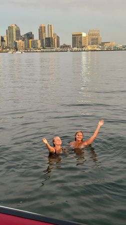 Two swimmers waving in a calm harbor at sunset, San Diego skyline and waterfront lights reflecting on the water.