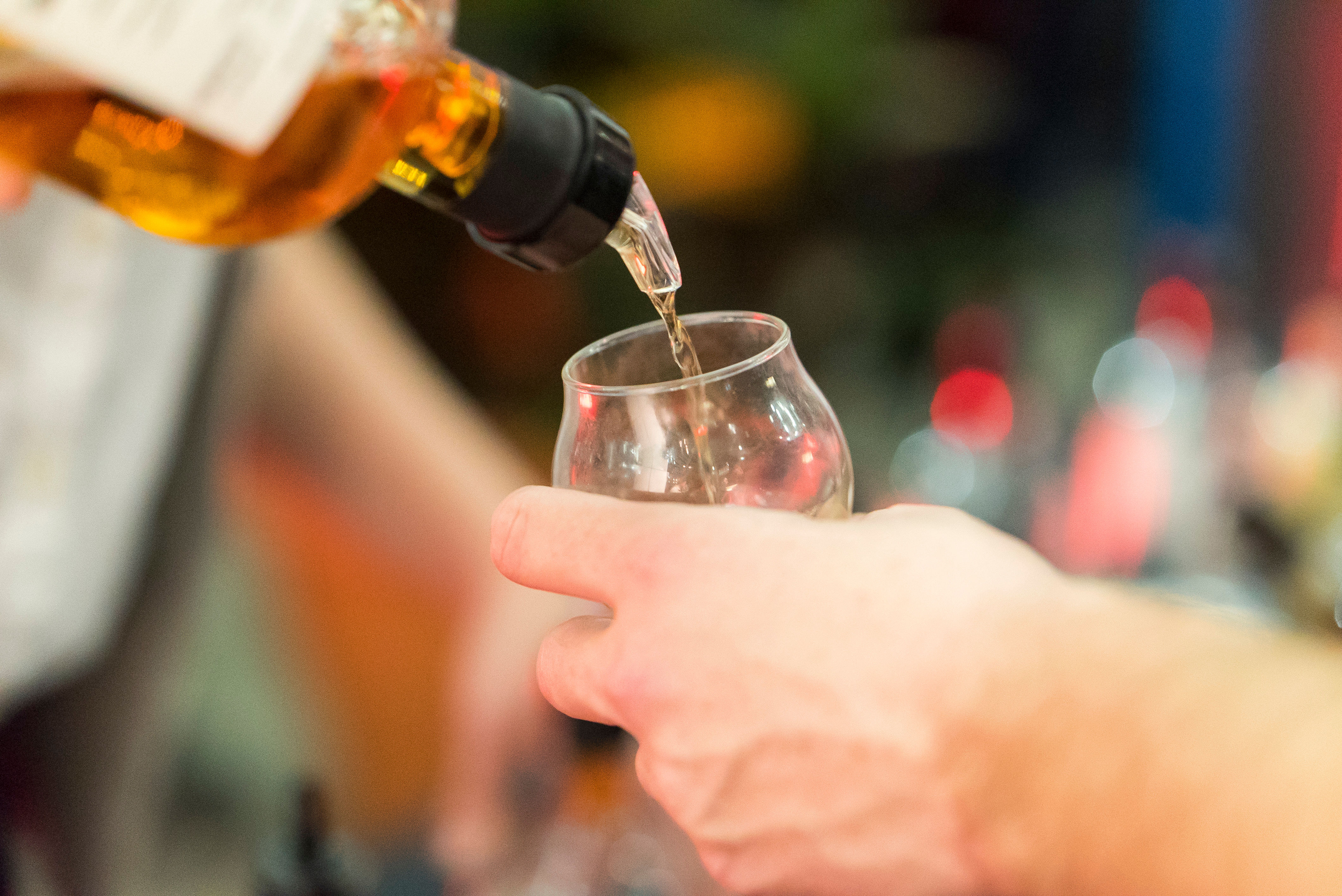 Close-up of amber whiskey being poured into a tulip tasting glass, hand-held pour in a lively bar setting with colorful bokeh background