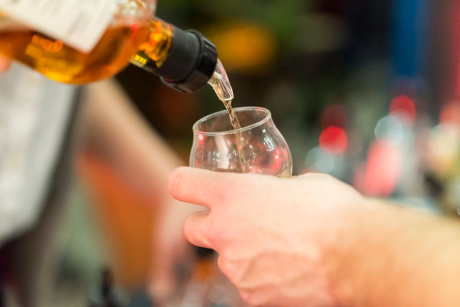 Close-up of amber whiskey being poured into a tulip tasting glass, hand-held pour in a lively bar setting with colorful bokeh background