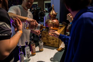 Pouring whiskey samples at an indoor distillery tasting booth with copper still, bottles, and visitors