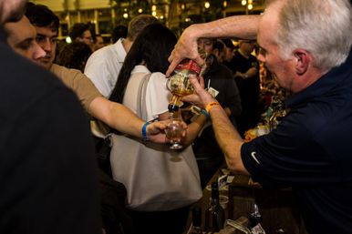 Server pouring a whiskey sample into a tasting glass at a crowded indoor craft spirits tasting event