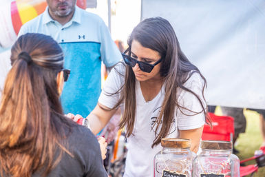 Sunglass-clad vendor pouring a drink sample from a can for a customer at a sunny outdoor market stall with glass jars on the table.