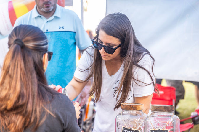 Sunglass-clad vendor pouring a drink sample from a can for a customer at a sunny outdoor market stall with glass jars on the table.