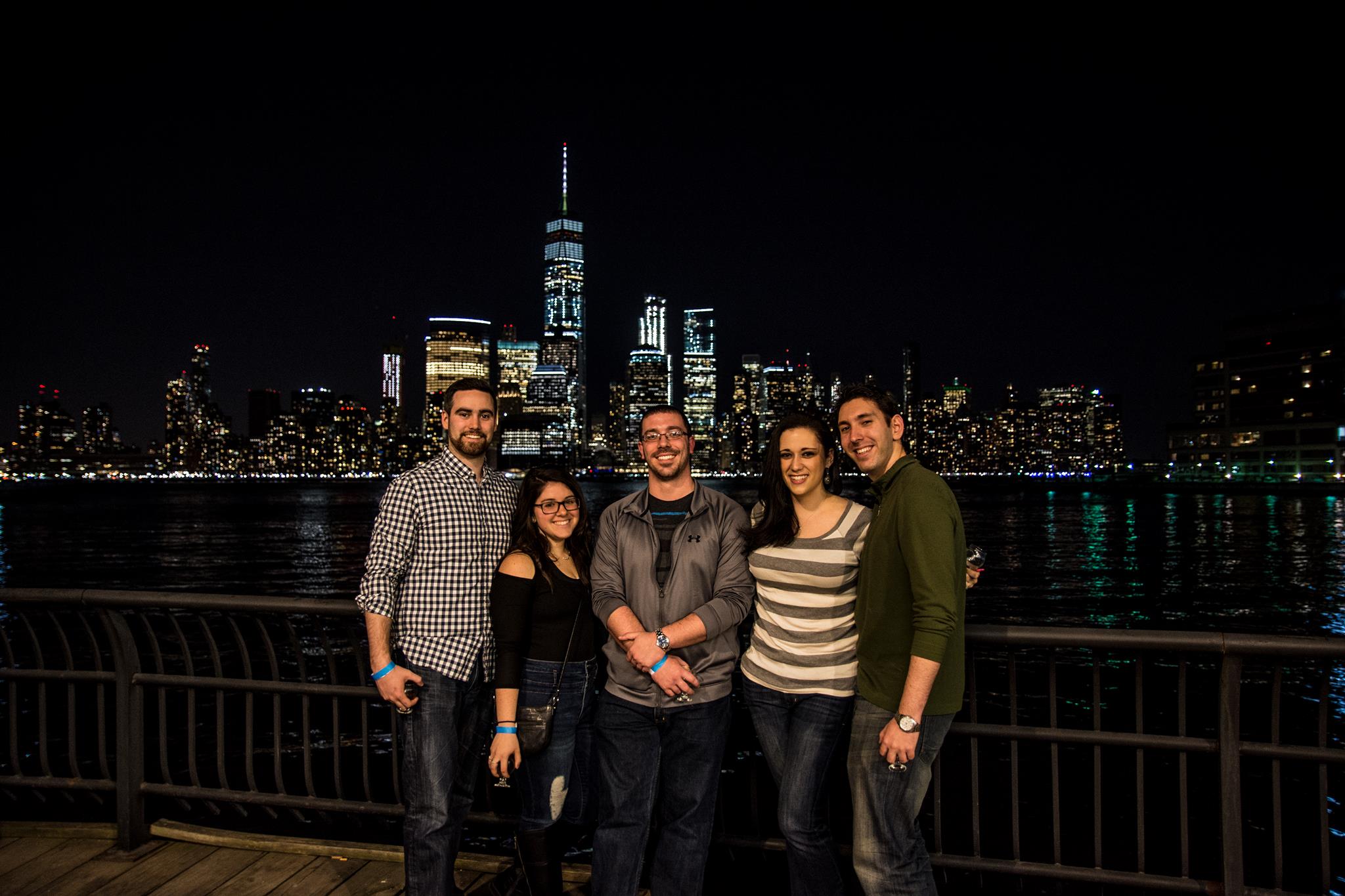 Smiling group of five posing on a waterfront promenade at night with the illuminated Manhattan skyline and One World Trade Center reflected in the Hudson River.