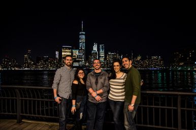 Smiling group of five posing on a waterfront promenade at night with the illuminated Manhattan skyline and One World Trade Center reflected in the Hudson River.