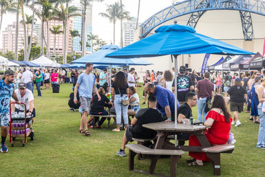 Crowded outdoor food and drink festival in a sunny urban park with palm trees, blue umbrellas over picnic tables, and rows of vendor tents and people milling around.
