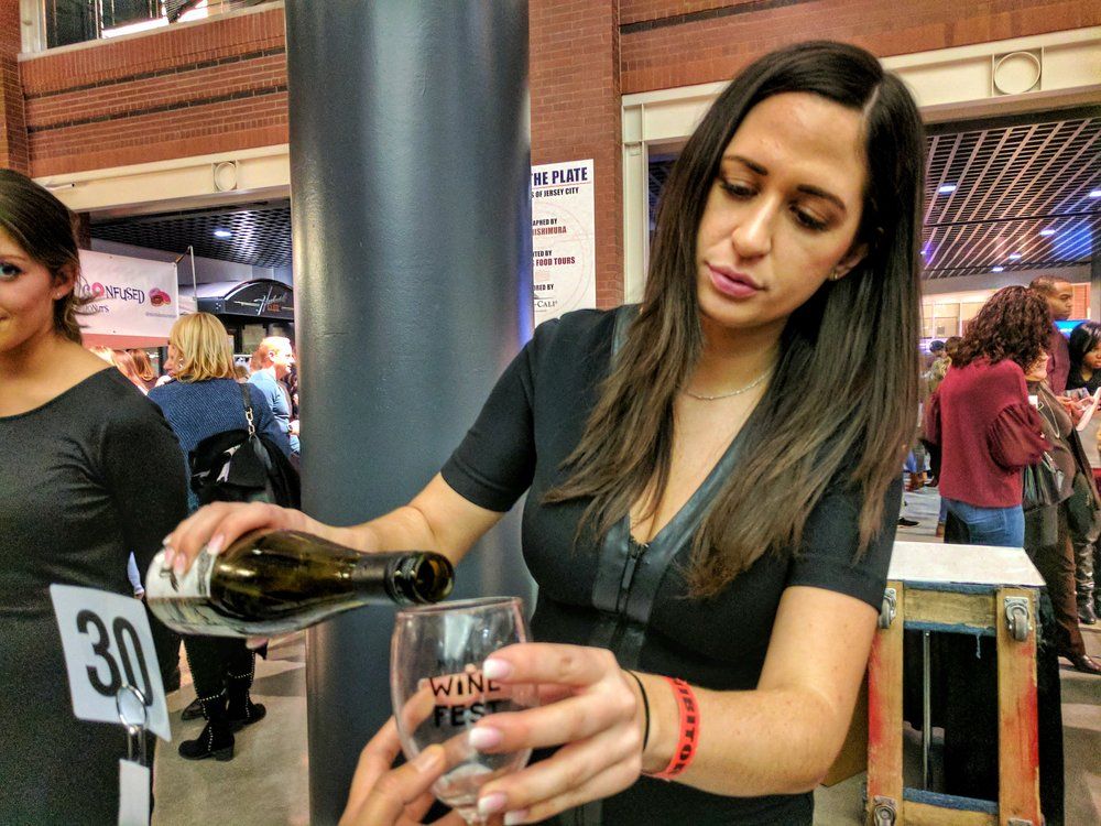 Server pouring wine into a tasting glass at a lively indoor wine festival booth with a bustling crowd