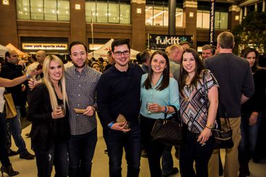 Five friends smiling and posing at a crowded indoor NYC food-and-drink festival, holding small plates and wine glasses with vendor tents and a lively crowd in the background.