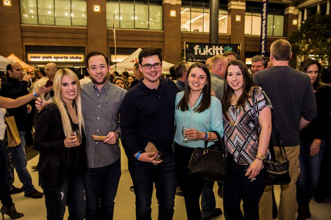 Five friends smiling and posing at a crowded indoor NYC food-and-drink festival, holding small plates and wine glasses with vendor tents and a lively crowd in the background.