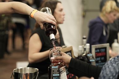 Hand pouring red wine into a tasting glass at an indoor wine-tasting event with blurred crowd and tasting table in the background.