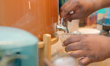 Hands operating a spigot on a large beverage dispenser, pouring a pale orange iced drink into a small clear plastic cup at a refreshment station.