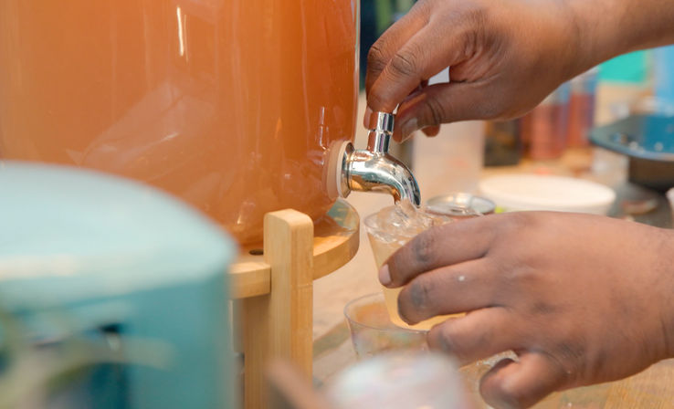Hands operating a spigot on a large beverage dispenser, pouring a pale orange iced drink into a small clear plastic cup at a refreshment station.