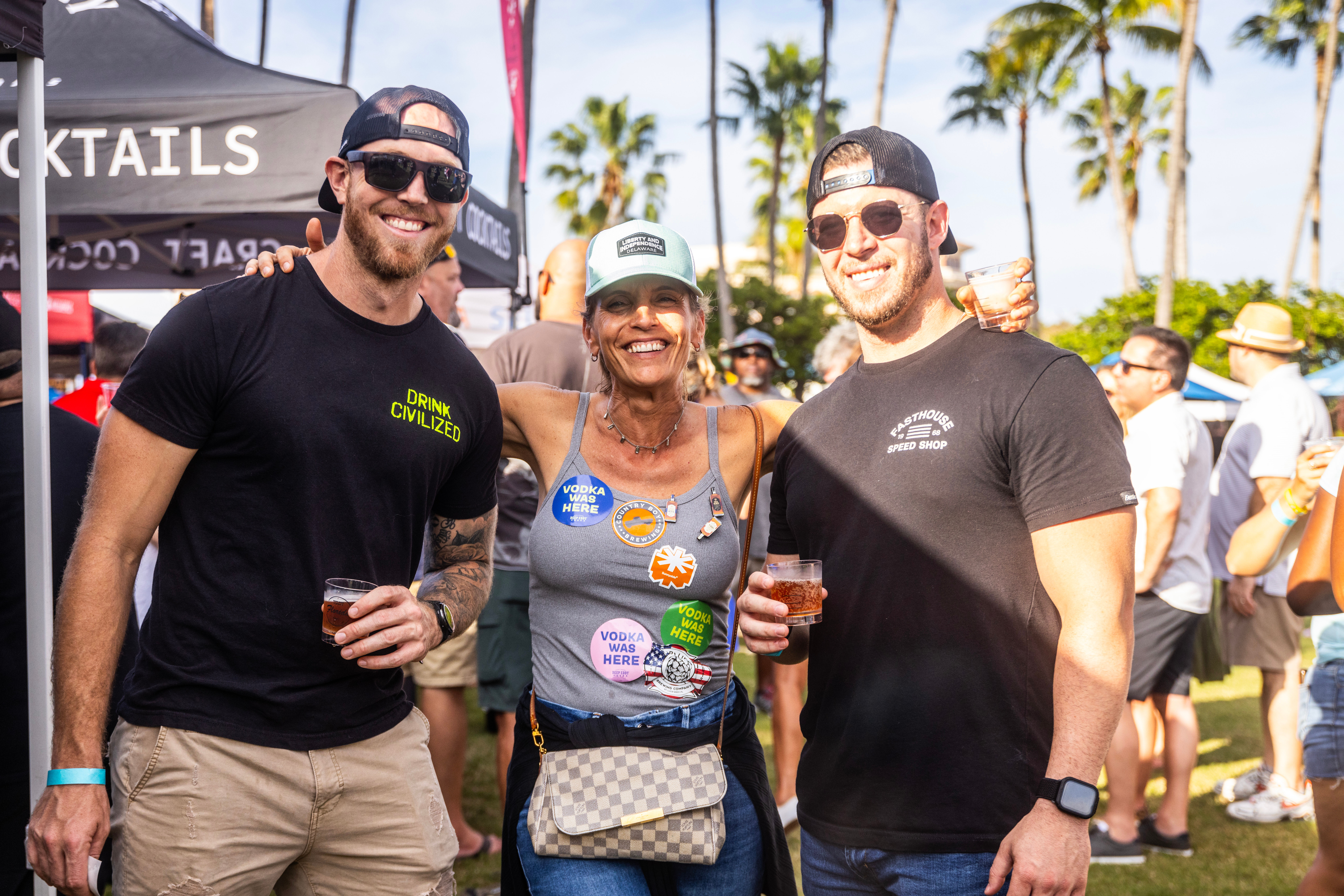 Three smiling adults in baseball caps and sunglasses holding drinks at a sunny outdoor beer and cocktail festival with palm trees and vendor tents in the background.