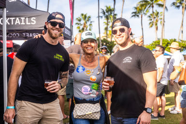 Three smiling adults in baseball caps and sunglasses holding drinks at a sunny outdoor beer and cocktail festival with palm trees and vendor tents in the background.