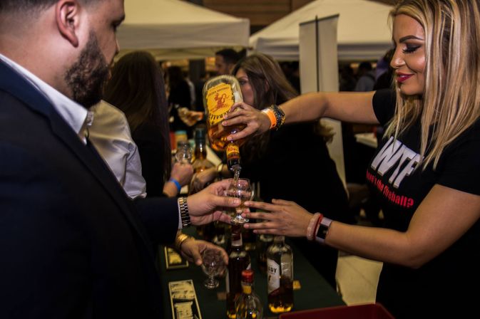 Smiling woman pours a whiskey sample into a small tasting glass for an attendee at a crowded indoor spirits tasting booth.
