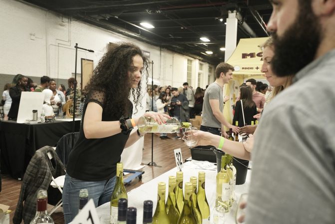 Woman pouring white wine for an attendee at a crowded indoor wine tasting event, bottles lined up on a vendor table in a busy expo hall