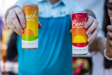 Hands holding two bright canned whiskey cocktails, mango lemonade and watermelon-peach flavors, summer-ready design.