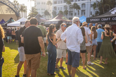 Sunny outdoor craft beer festival in a coastal park — crowds lined up at brewery and craft tents on a grassy lawn with palm trees and white beachfront buildings in the background.