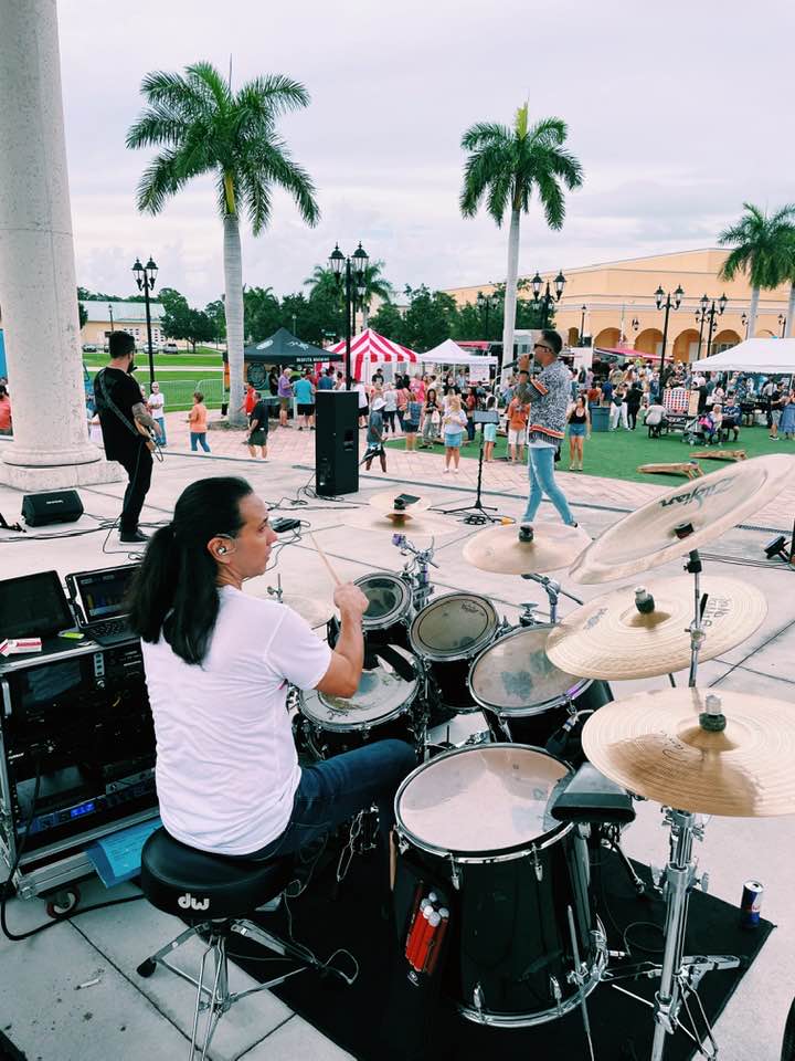Drummer behind a black drum kit performing on an outdoor stage at a palm-tree–lined music festival with vendor tents and a crowd in a tropical plaza