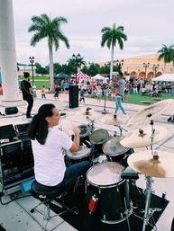 Drummer's view of a live band on an outdoor stage at a palm-lined community festival with vendor tents and a lively crowd.