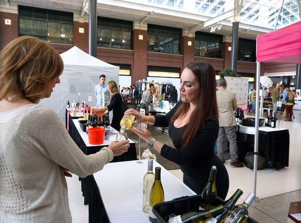 Server pouring white wine into a guest’s glass at an indoor wine-tasting event in a bright atrium-style market or convention hall, with wine bottles and vendor booths on tables