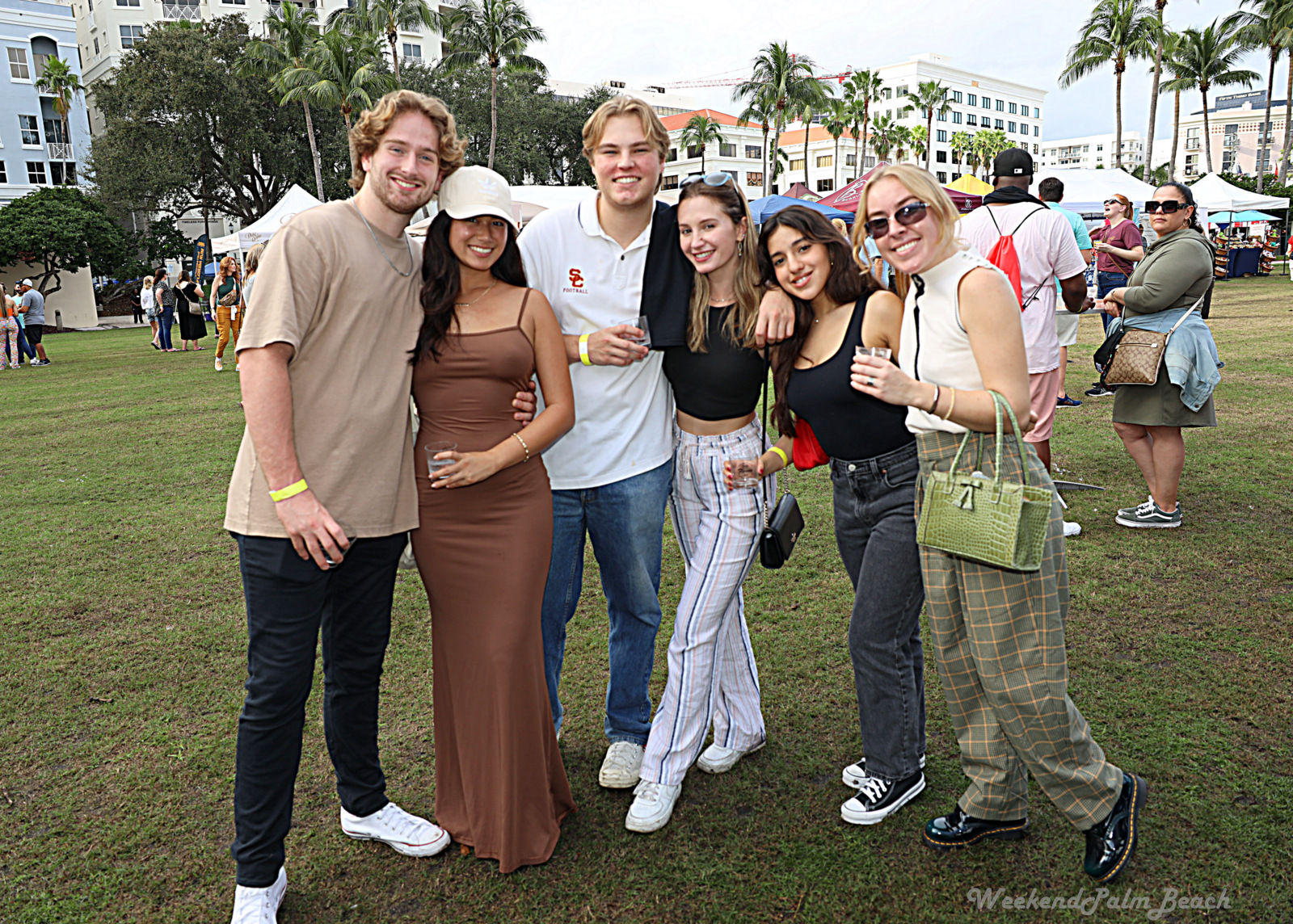 Six friends smiling and posing at an outdoor Palm Beach park festival with palm trees, vendor tents and city buildings in the background; casual outfits and drinks, relaxed group photo.