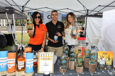 Cheerful vendors at an outdoor market booth holding bottles for a spirits tasting, with tequila and whiskey bottles, colorful packaging and signage under a white canopy.