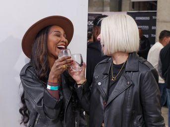 Two friends in leather jackets clinking stemless wine glasses and laughing at an indoor social event, one wearing a brown hat and the other with a short platinum bob.