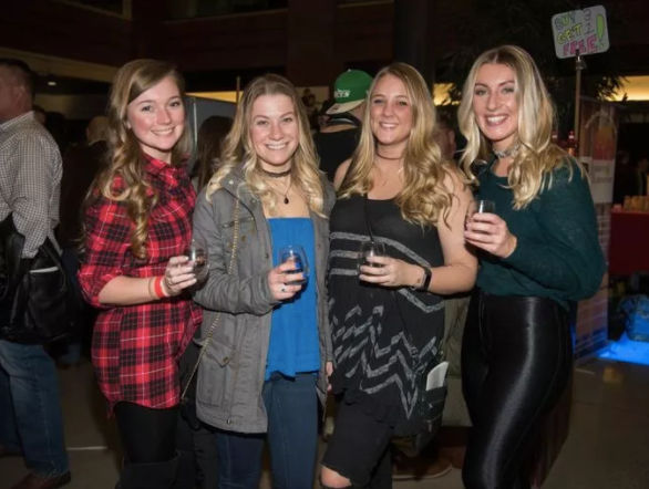 Four smiling women holding drinks and posing at an indoor nightlife event — casual outfits and a lively night-out vibe