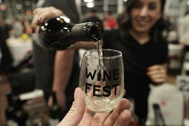 Close-up of white wine being poured from a bottle into a stemless glass labeled 'WINE FEST' at a busy indoor wine-tasting event.