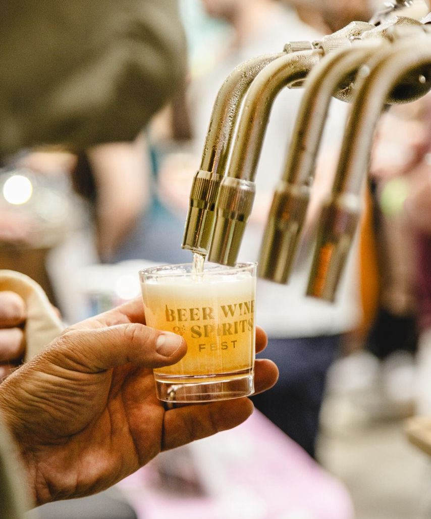 Close-up of a hand holding a sample glass labeled "Beer Wine & Spirits Fest" as golden beer pours from stainless steel draft taps at a busy craft beer tasting festival