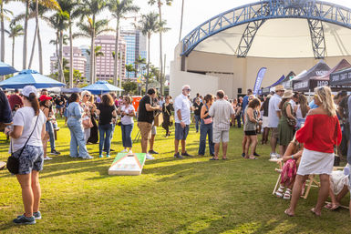 Sunny outdoor festival at a palm‑lined amphitheater with crowds, vendor tents, and a cornhole game on the grassy lawn
