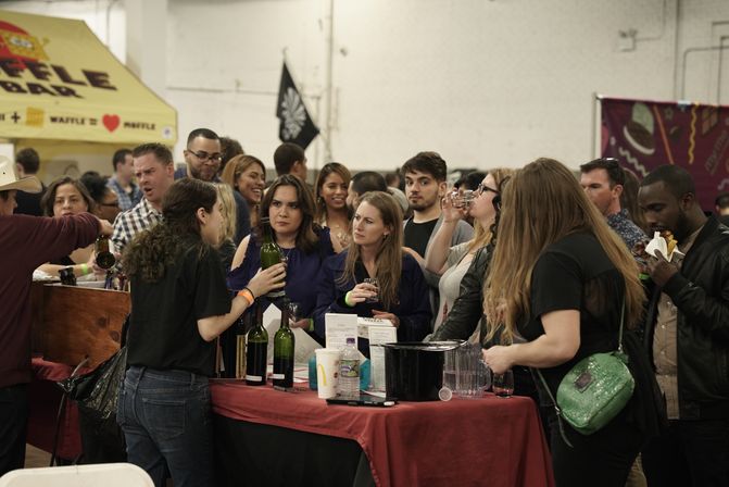 Busy indoor wine-tasting festival crowd sampling bottles and glasses at a vendor booth with a red tablecloth, pitchers, and food tents in the background.