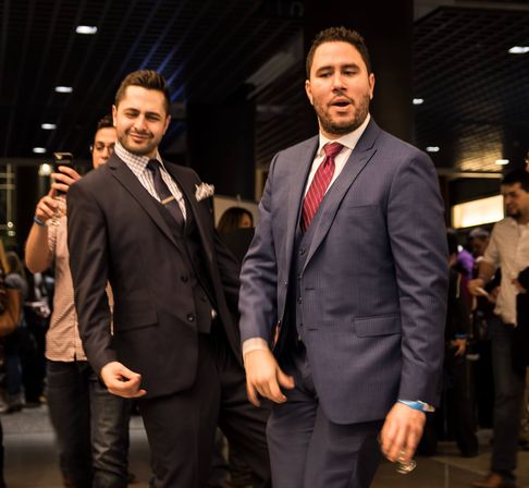 Two men in suits striking playful poses at a crowded indoor networking event, guests and photographers in the background.
