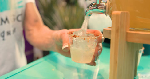 Hand holding a small cup rimmed with chili powder as a dispenser pours a cold citrus drink sample at a colorful market stall