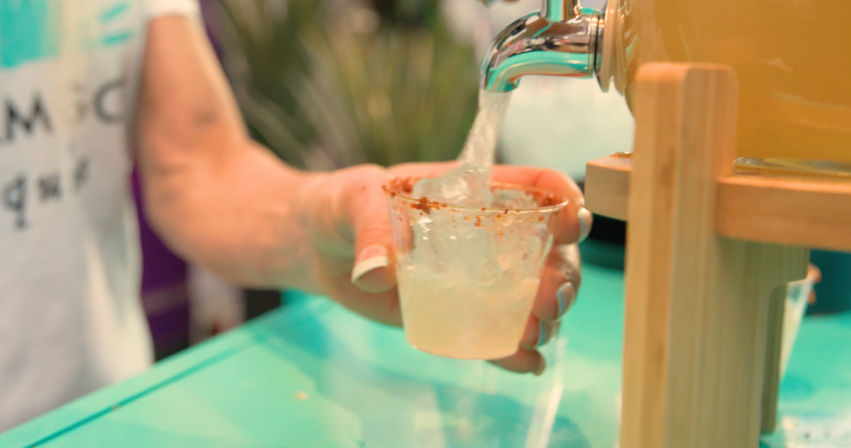 Hand holding a small cup rimmed with chili powder as a dispenser pours a cold citrus drink sample at a colorful market stall