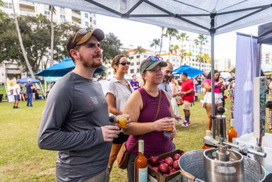 People sampling craft beverages at an outdoor tasting tent in a palm-tree park festival, holding plastic cups near bottles, apples and a drink press with crowds and city buildings in the background.