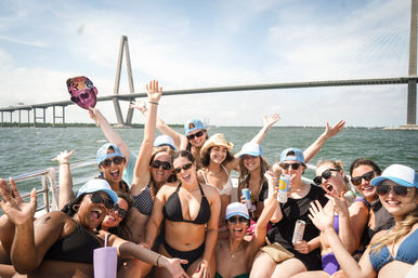 Cheerful group of women on a sunny boat party by a cable-stayed bridge over a coastal bay, wearing swimsuits and caps, waving and holding drinks.