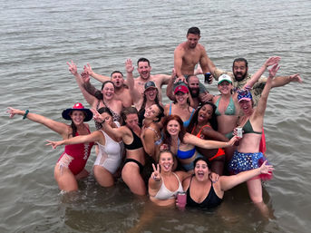 Cheerful mixed group of adults in swimsuits posing and cheering waist-deep in shallow coastal water, holding drinks on an overcast summer outing.