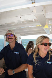Smiling male and female crew in navy polo shirts and sunglasses enjoying a sunny boat ride on choppy coastal ocean waters.