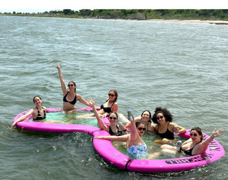 Eight friends in swimsuits smiling and waving from two connected pink inflatable floats on a sunny coastal bay, holding canned drinks with a tree-lined shoreline in the background.