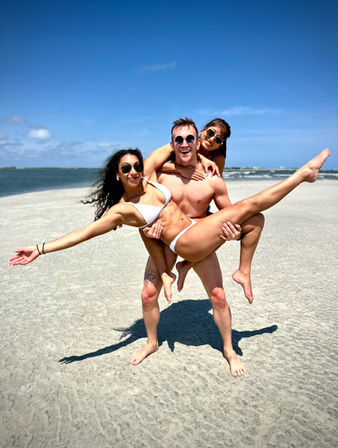 Three friends laughing on a sunny sandy beach — shirtless man carrying two women in bikinis, all wearing sunglasses, with ocean waves and a clear blue sky in the background.