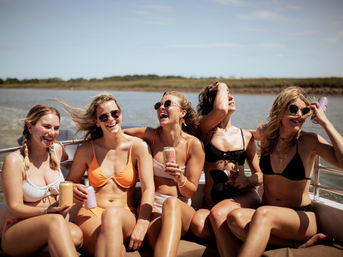 Five women in bikinis and sunglasses laughing and holding canned drinks on a motorboat cruising along a coastal marsh on a sunny summer day.