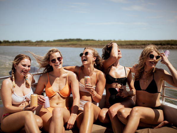 Five women in bikinis and sunglasses laughing and holding canned drinks on a motorboat cruising along a coastal marsh on a sunny summer day.