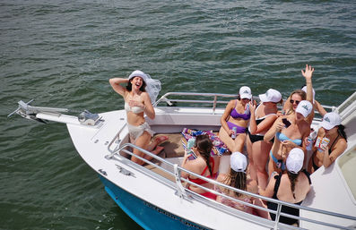 Bride-to-be in a white bikini and veil laughing at the bow of a boat while friends in swimsuits and matching white caps cheer, sip drinks, and take photos on open water.
